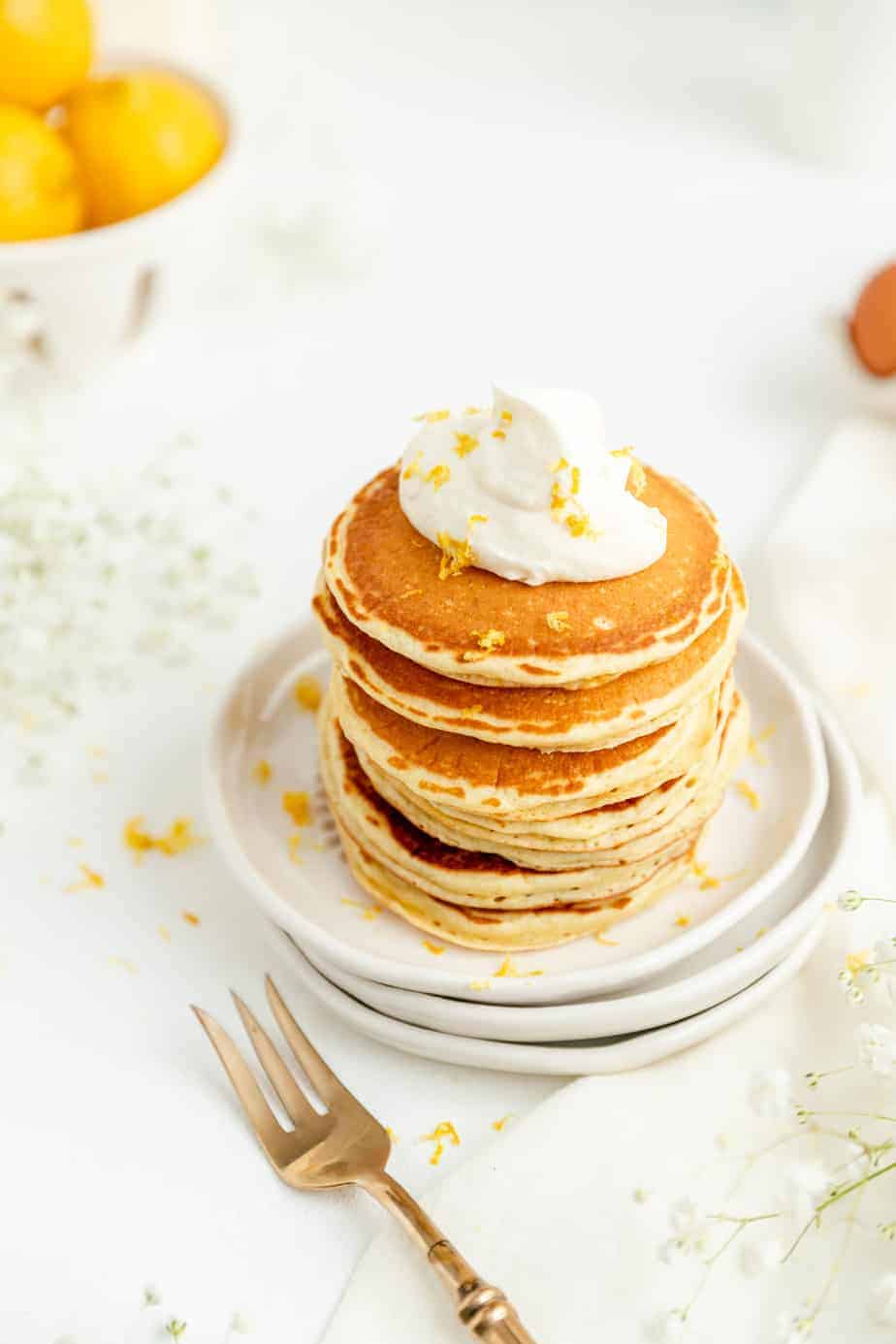 45° shot of lemon pancakes stacked up with whip cream and lemons in bowl in back.