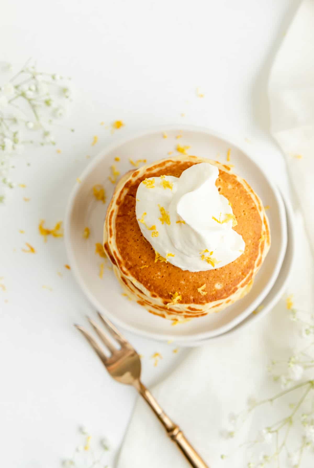 Overhead of lemon pancakes with whipped cream and zest with fork and flowers on table.