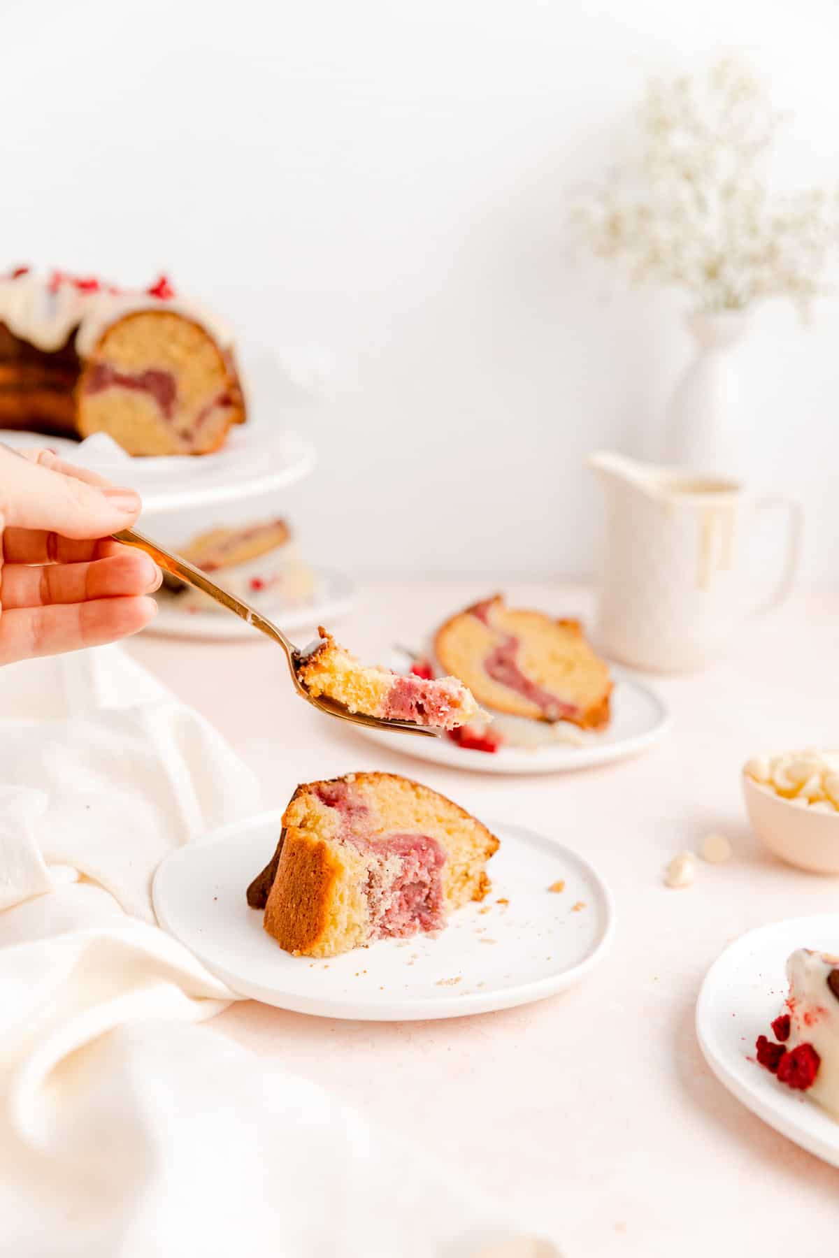 Hand holding bite of white chocolate raspberry Bundt cake on fork with plated slices on table.