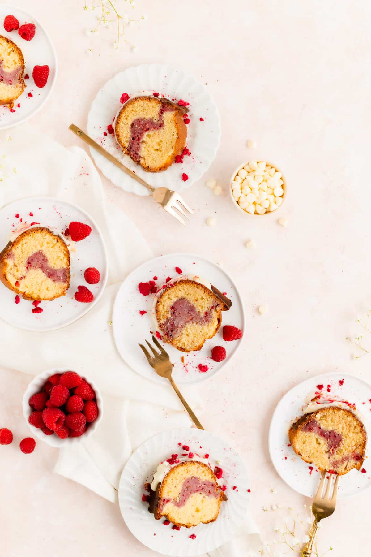 Six plated slices of white chocolate raspberry bundt cake with bowls of raspberries and chocolate chips.