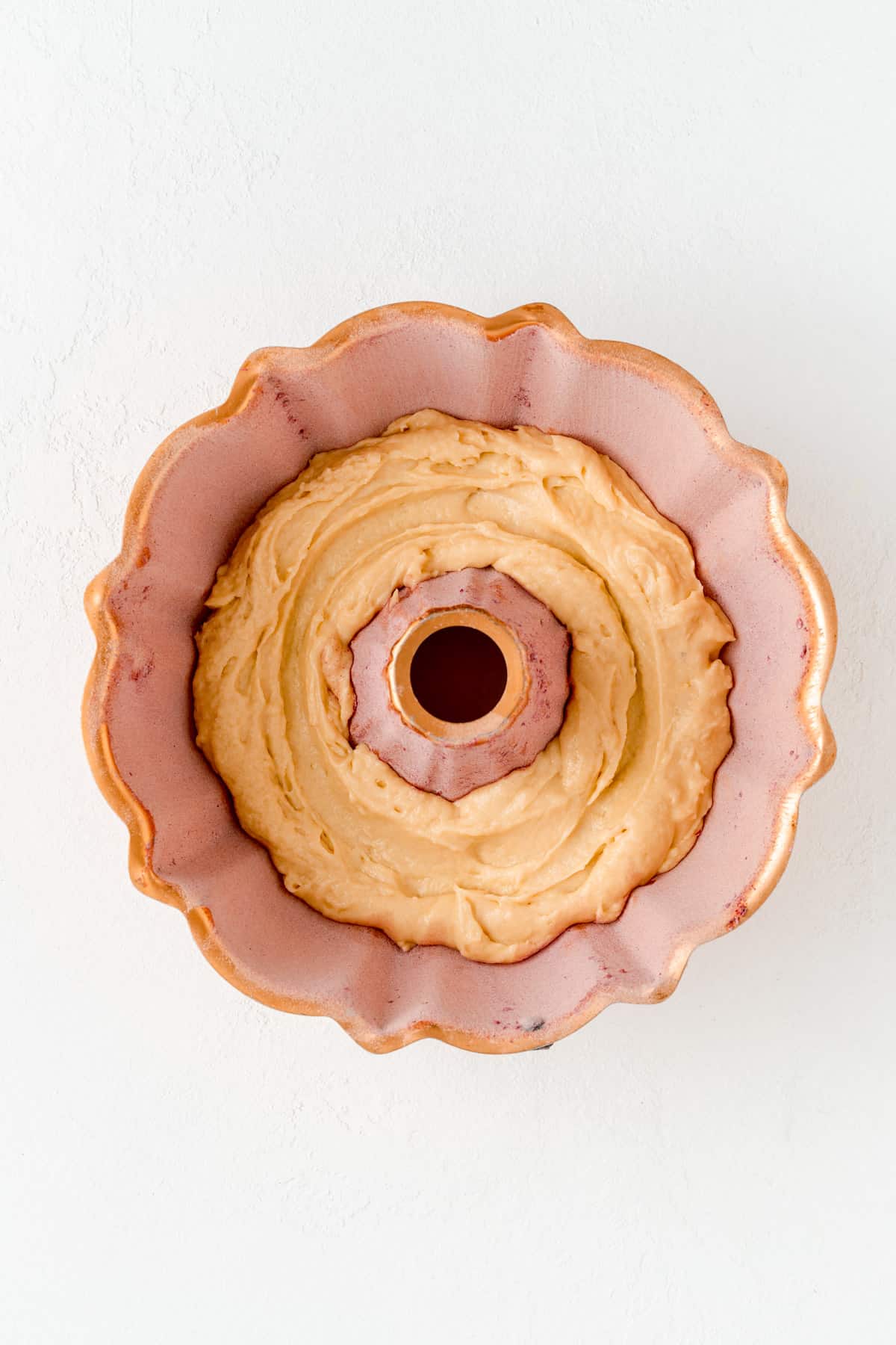 Copper Bundt pan with vanilla batter layer in bottom on white background.