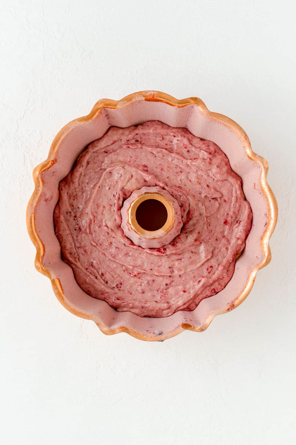 Copper Bundt pan with raspberry batter layer on white background.