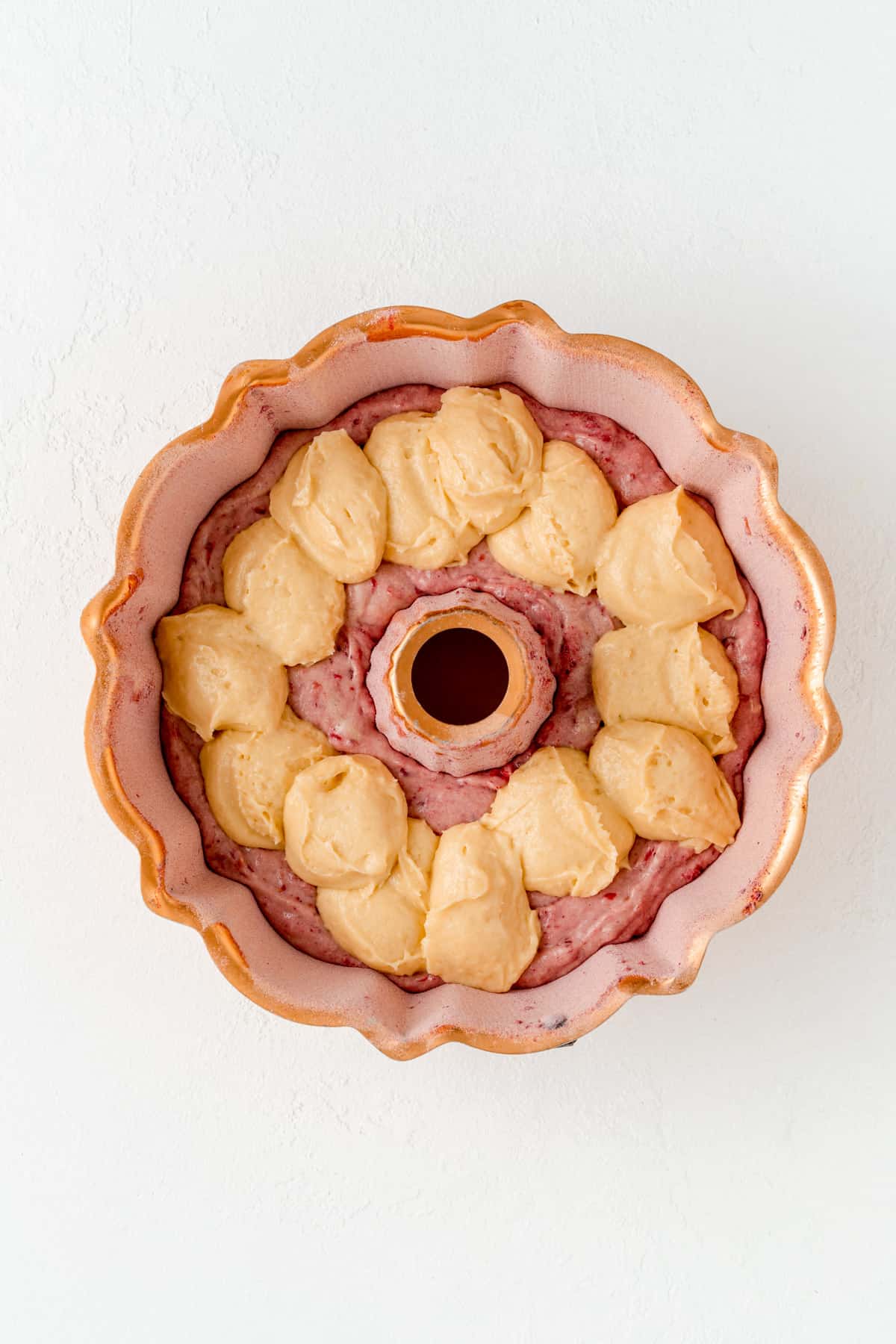 Copper Bundt pan topped with spoonfulls of vanilla batter overtop raspberry batter layer.