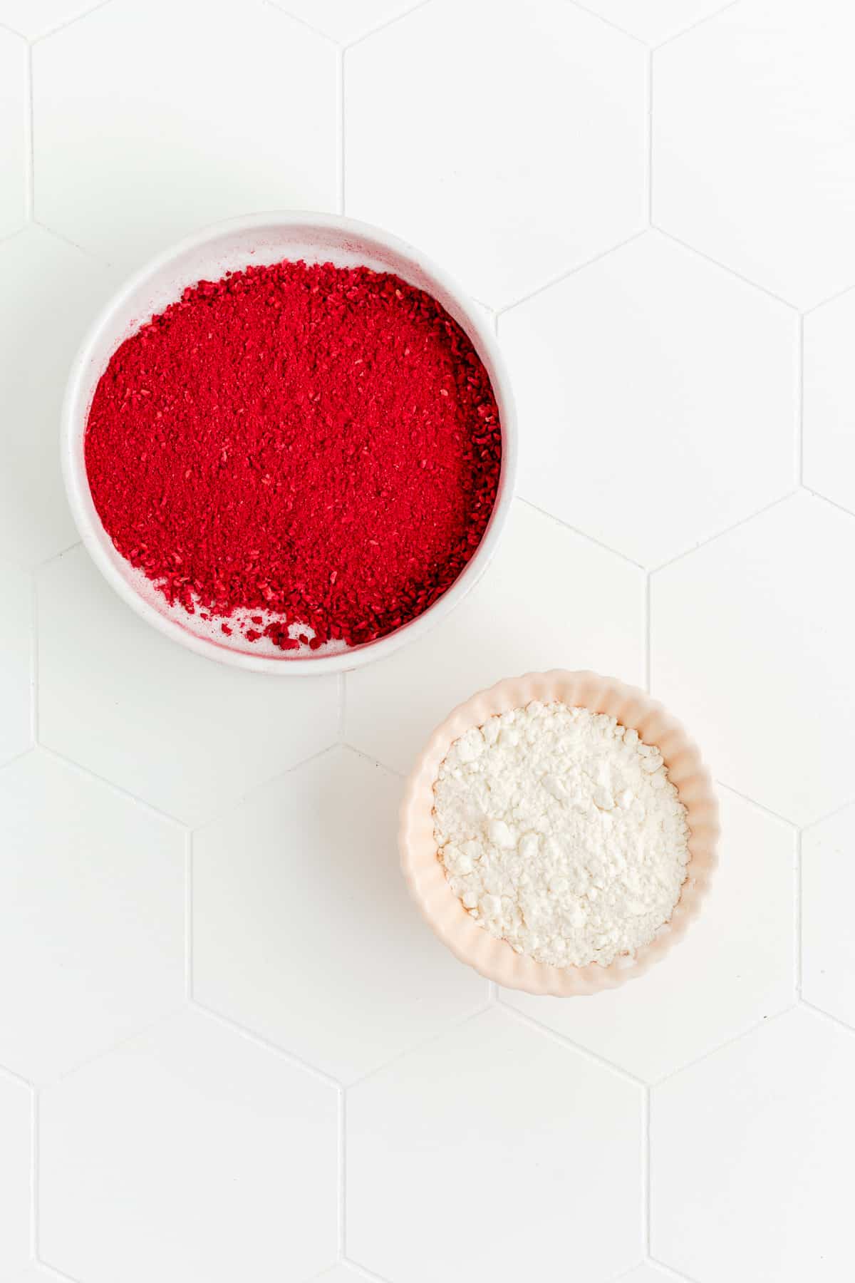 Crushed freeze dried raspberries and flour in individual bowls on white background.