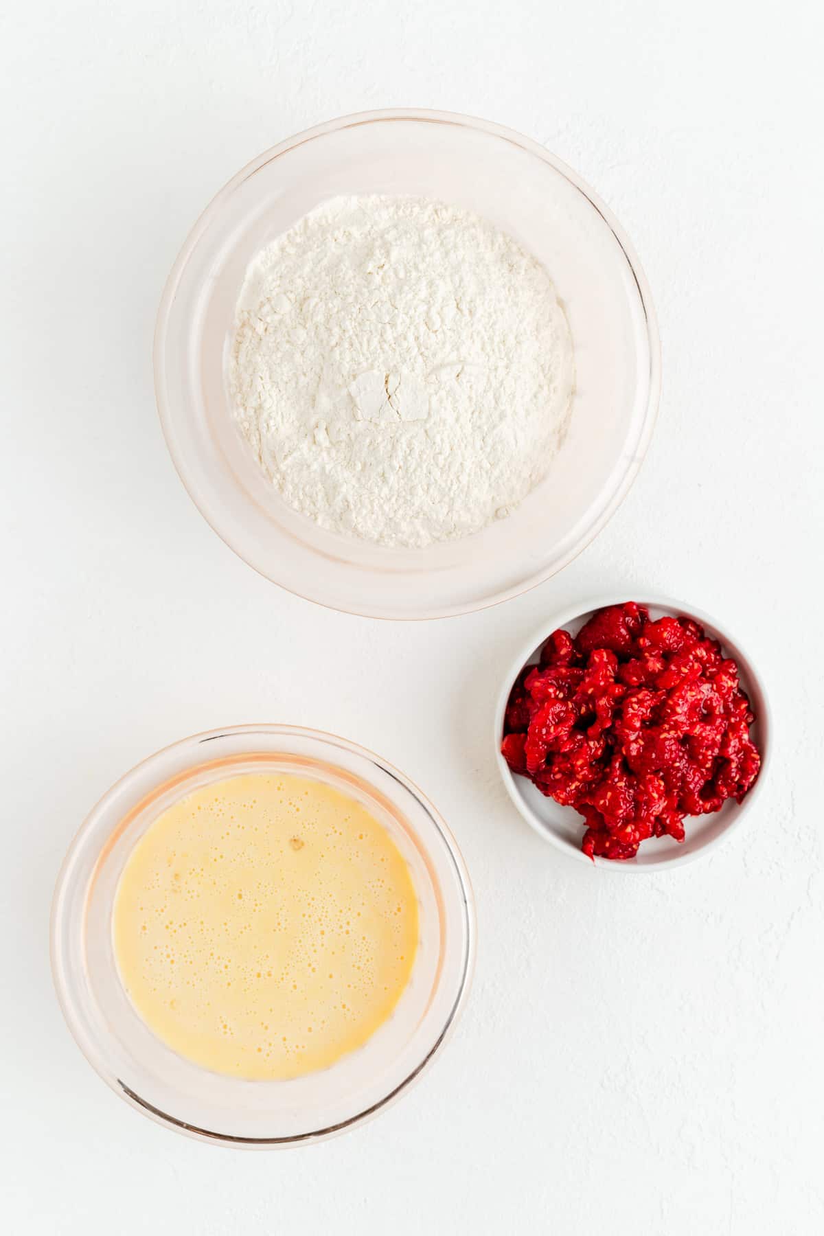 Dry ingredients, wet ingredients, and crushed fresh raspberries in their own bowls on white background.