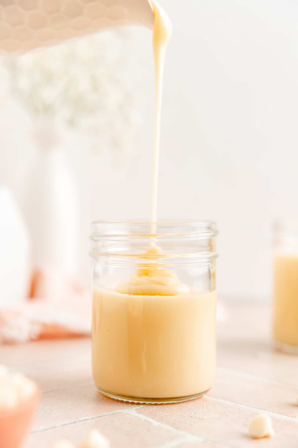 White chocolate sauce pouring from pitcher in to clear mason jar on brick surface.