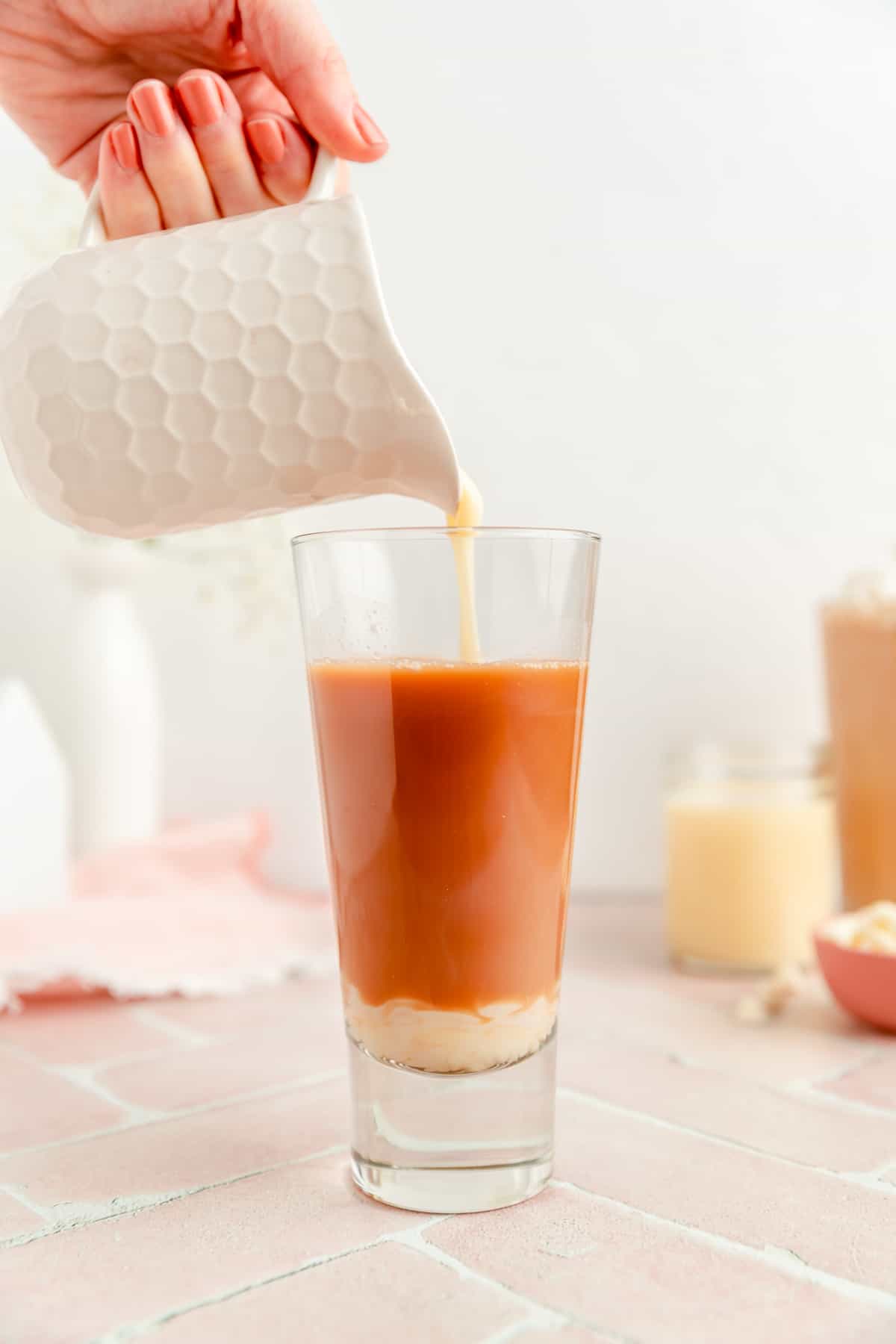 Hand holding white honeycomb pitcher pouring white chocolate sauce in tall glass of coffee.