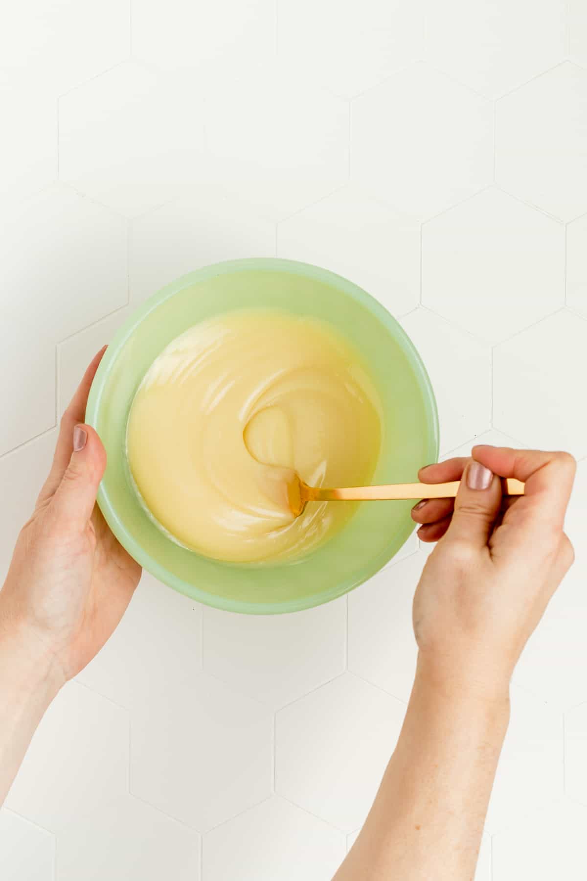 Hand holding green mixing bowl and stirring white chocolate sauce with gold fork.