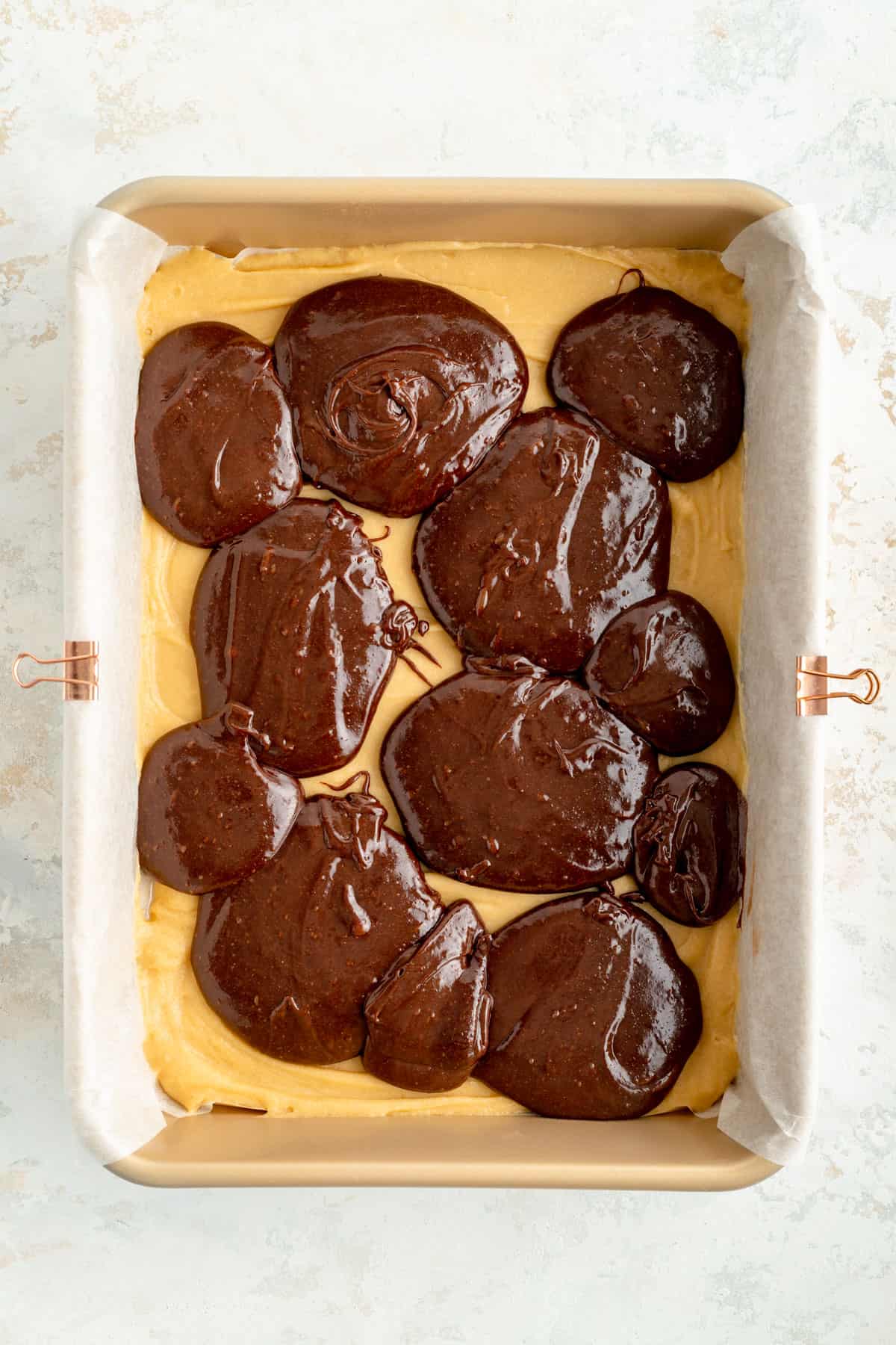 Blondie batter in Gold pan with large spoonfuls of brownie batter on top.
