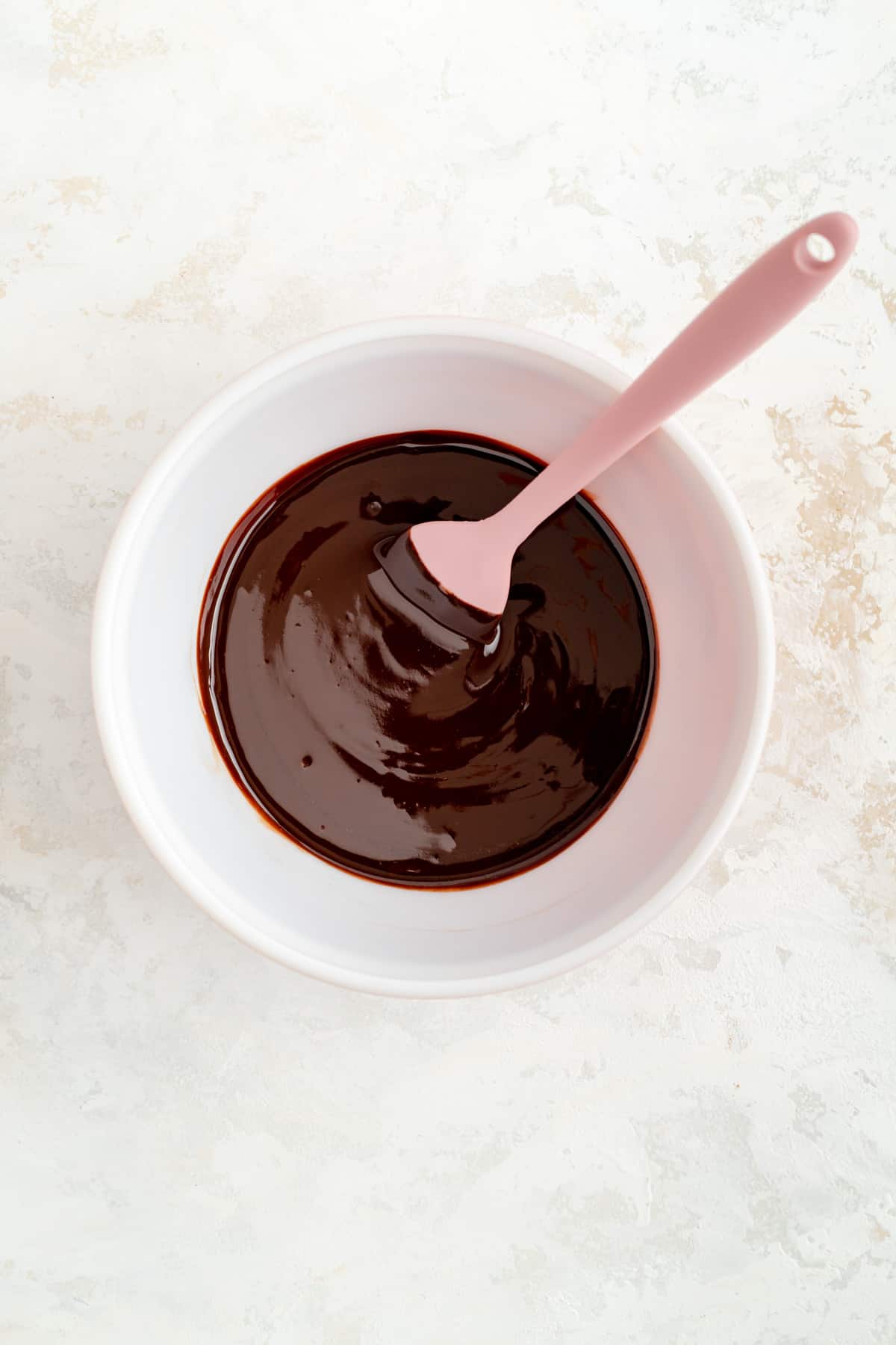 Melted chocolate and butter stirred together in a white bowl with a pink spatula.