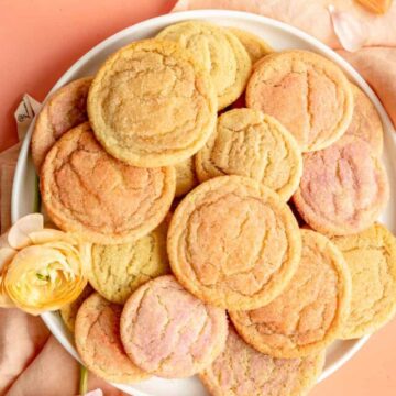 white plate piled with multi-colored sugar cookies on coral background with ranunculus on table.