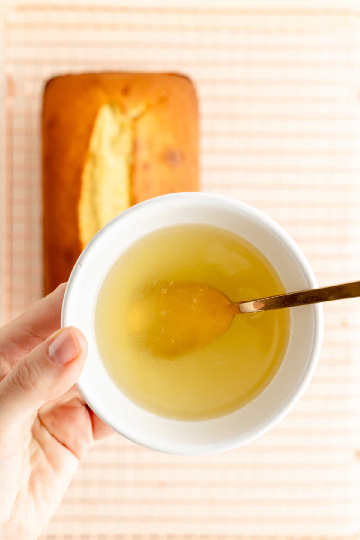 hand holding lemon syrup in a bowl over a cooling rack with a pound cake.