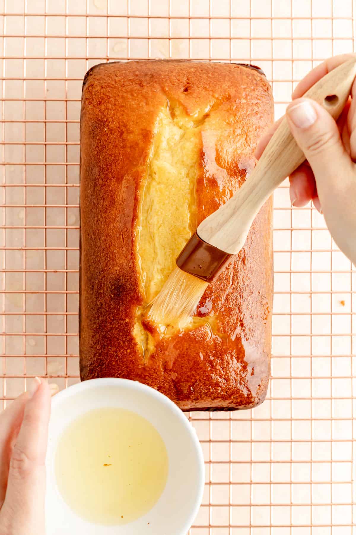 hand brushing a baked pound cake with lemon glaze from a bowl in the corner.