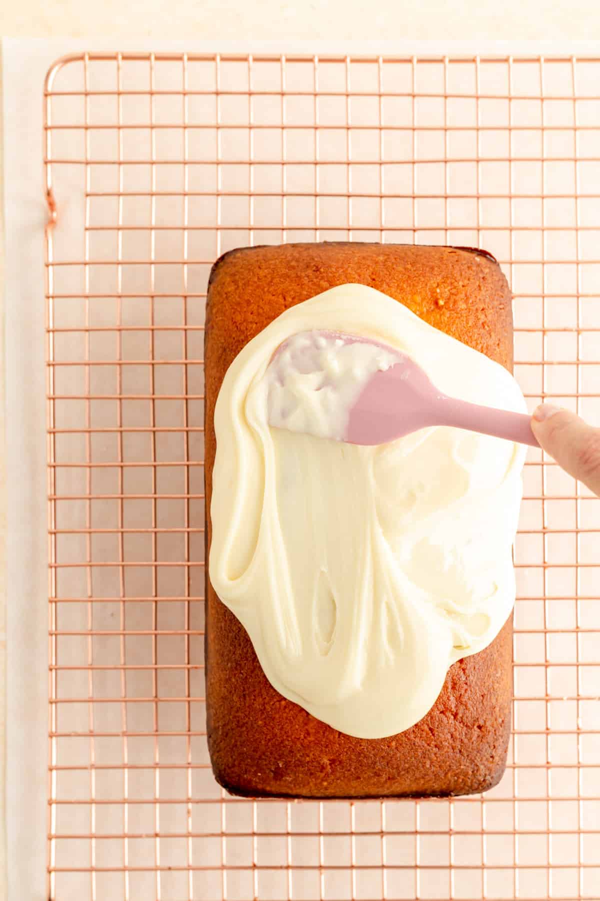 a hand with a pink spatula glazing a baked pound cake on a copper wire rack.