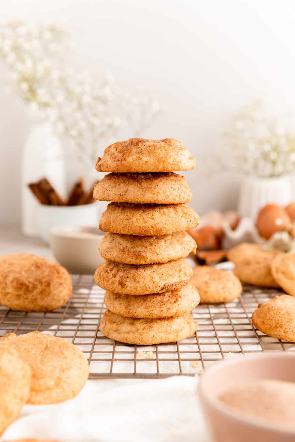 side shot of 7 stacked snickerdoodles on cooling rack with cookies and eggs in background.