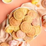 white plate piled with multi-colored sugar cookies on coral background with ranunculus on table.