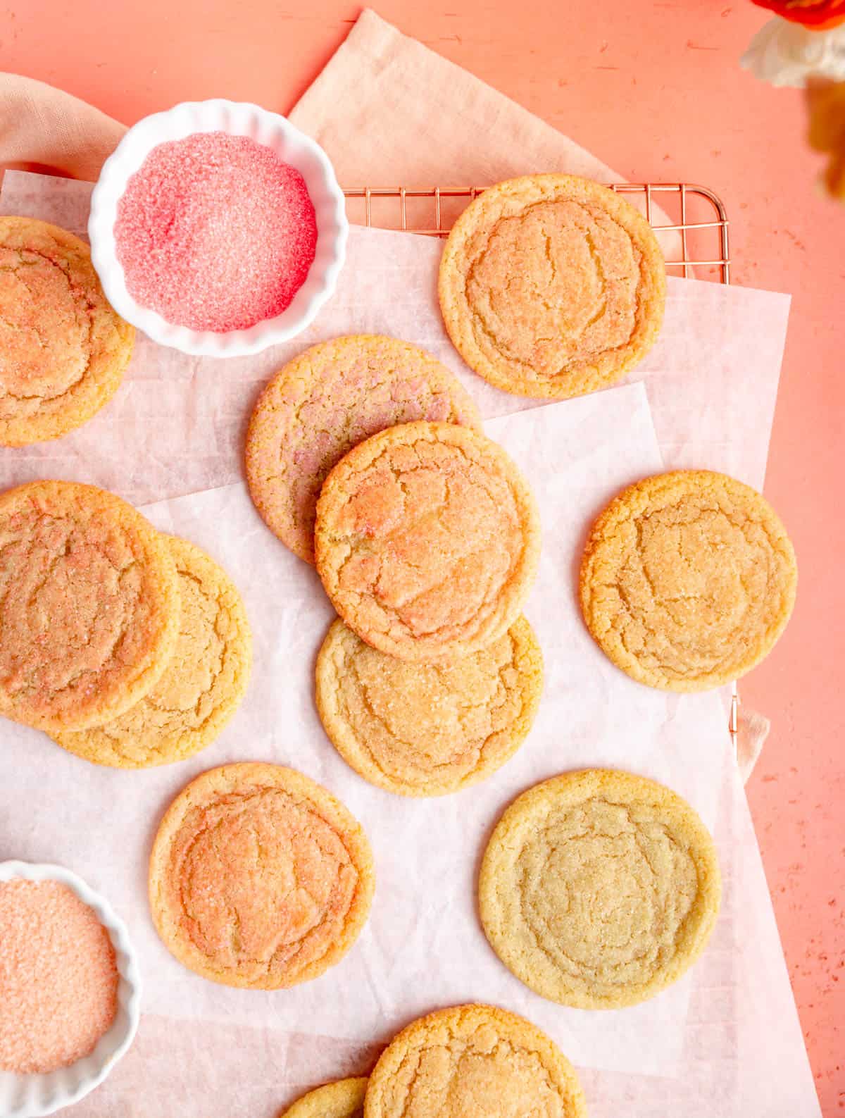 multi colored sugar cookies on parchment paper on coral background with bowls of colored sugar.