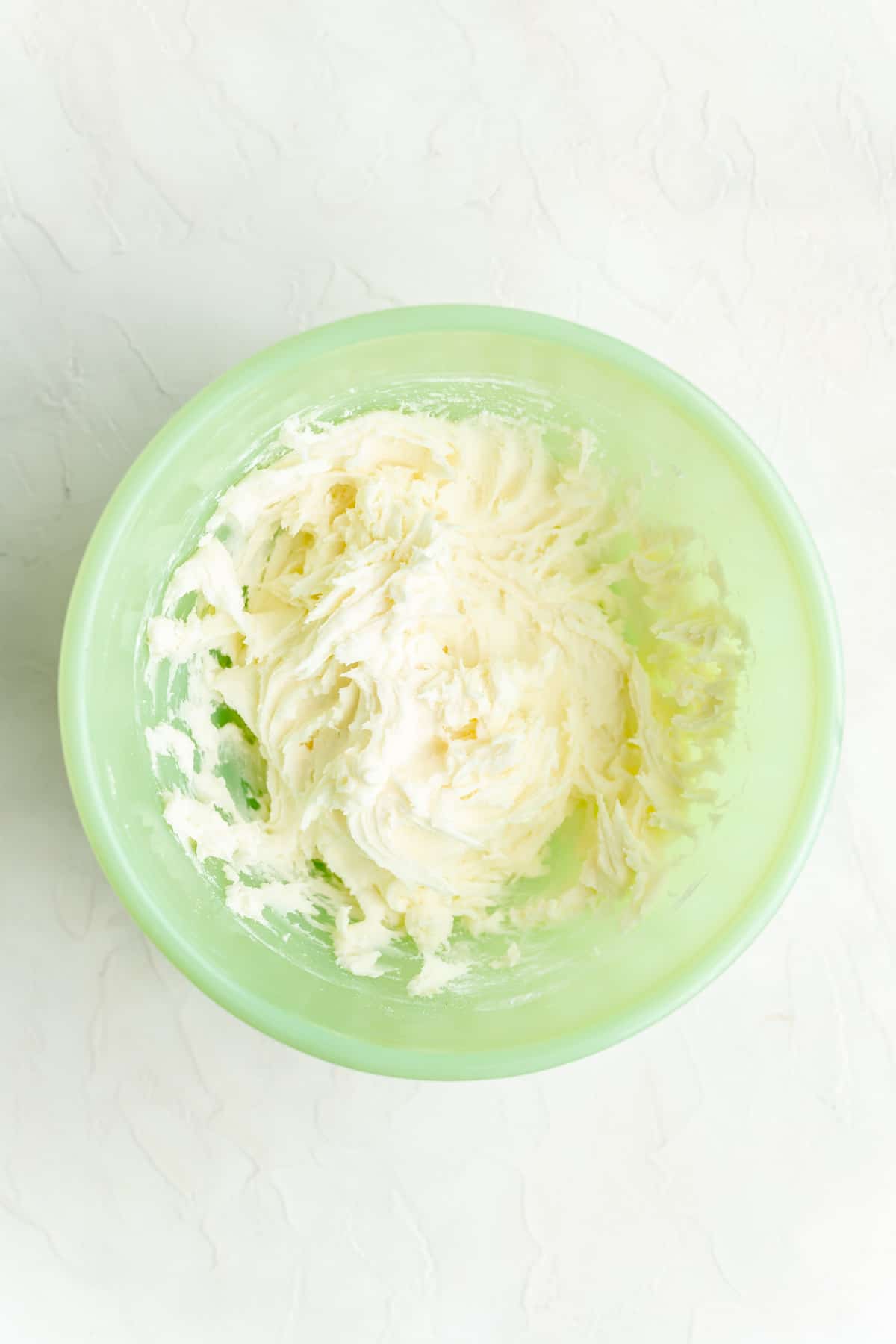 Partially mixed cream cheese icing in green mixing bowl on white background.