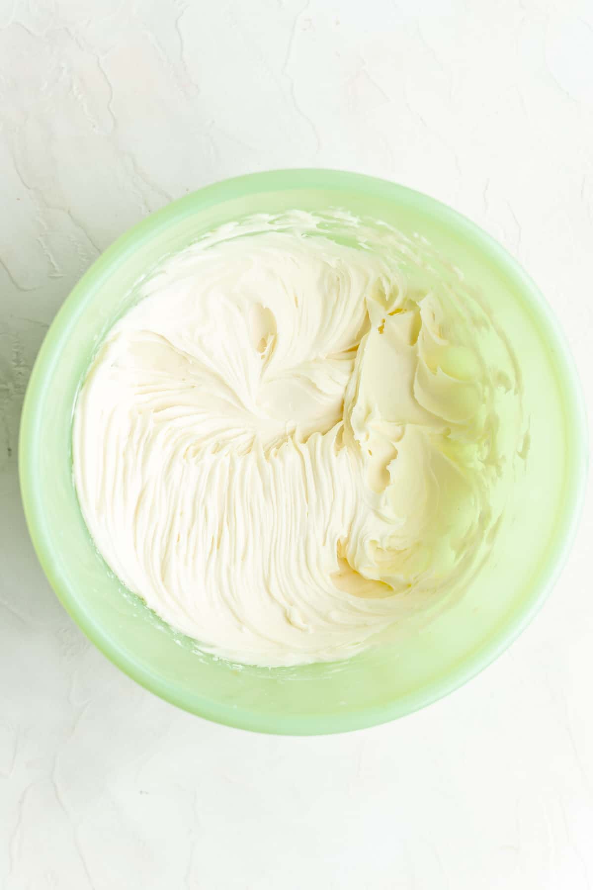 Fully mixed cream cheese icing in green mixing bowl on white background.