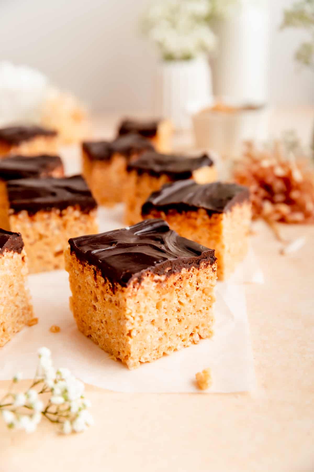 Side shot of chocolate peanut butter rice krispie treats scattered on white parchment with flowers around.