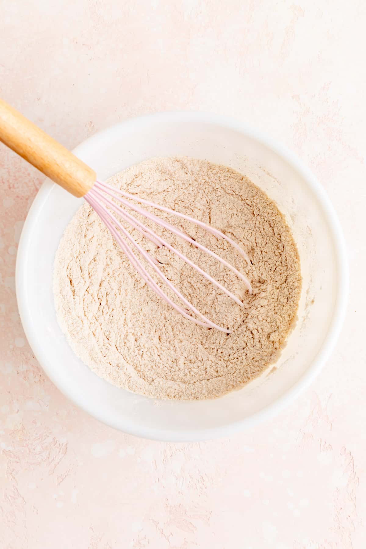 Dry ingredients for cinnamon pancakes in white ball with pink and wood whisk.