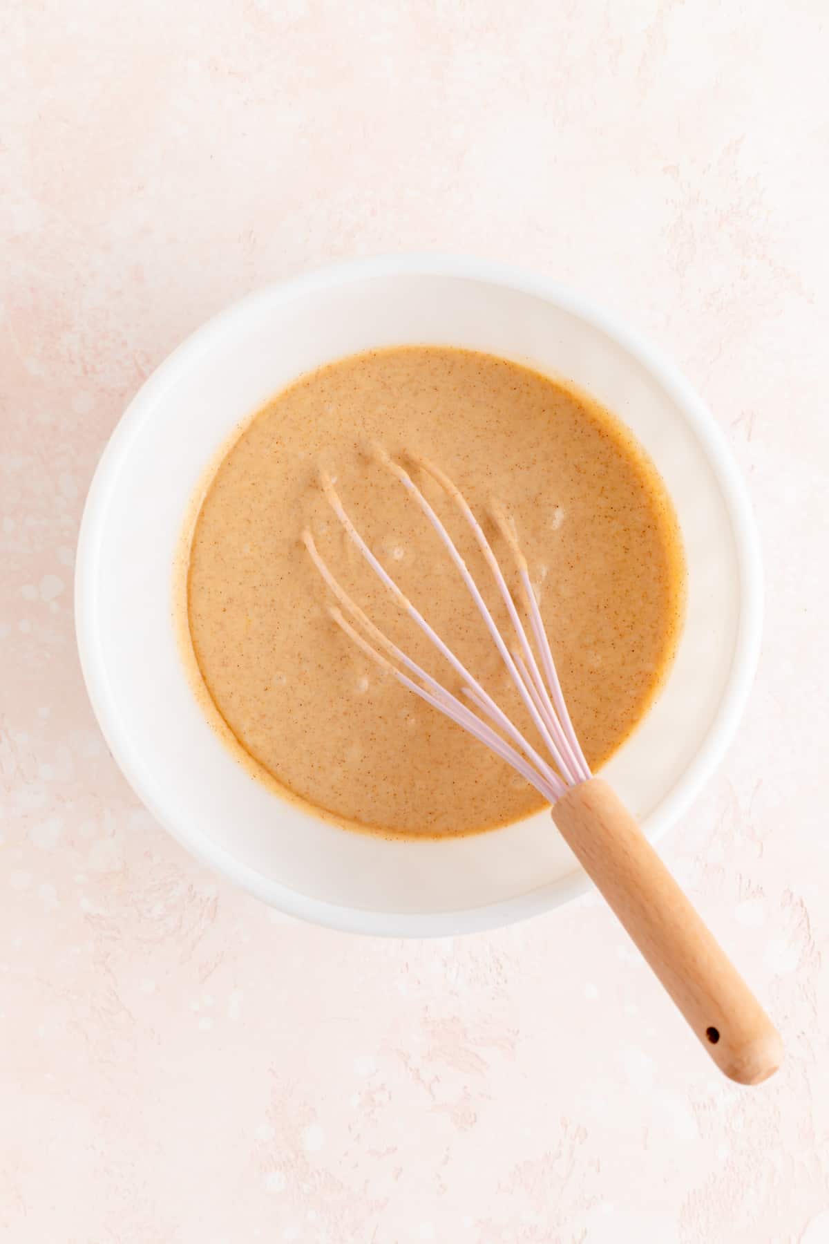 Cinnamon French toast pancake batter in white bowl with pink and wood whisk.