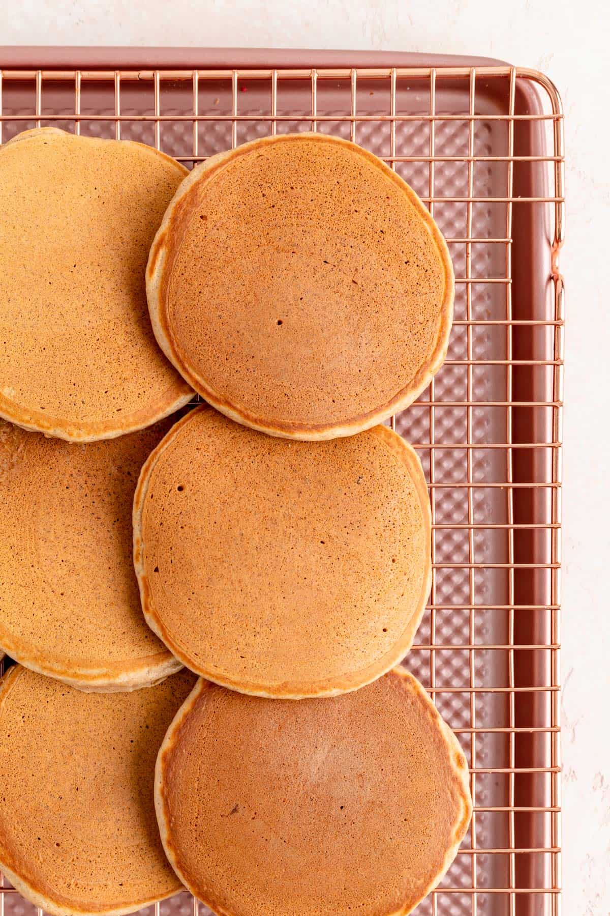 Cinnamon pancakes lined up on copper wire rack over pink baking sheet.