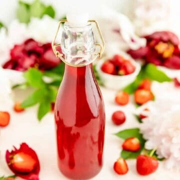 Close-up of red strawberry syrup in glass bottle with flowers and berries on table.