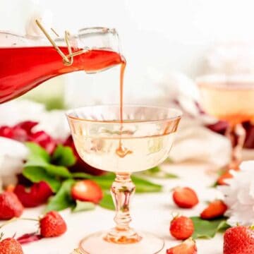 Glass bottle of strawberry syrup being poured into pick cocktail glass filled with Prosecco.