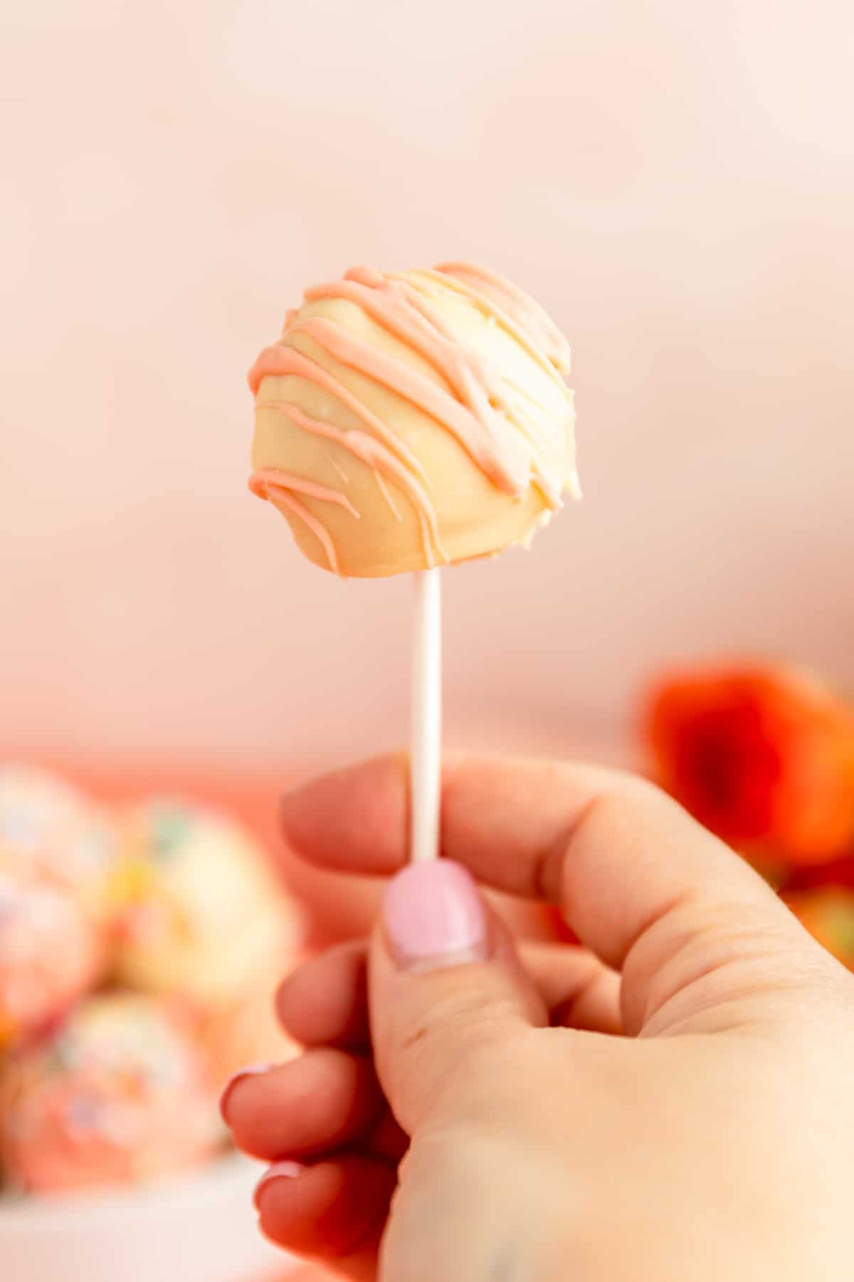 A hand holding a cookie dough lollipop decorated in white and pink chocolate..