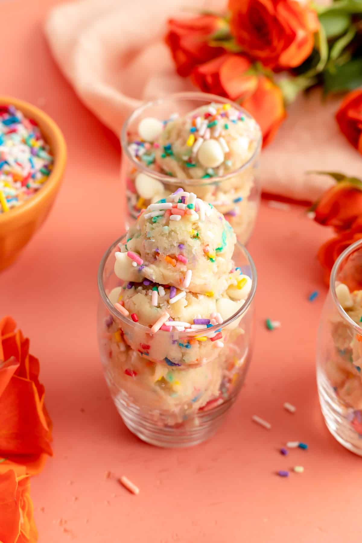 Three glasses filled with edible sugar cookie dough covered in sprinkles on pink background.