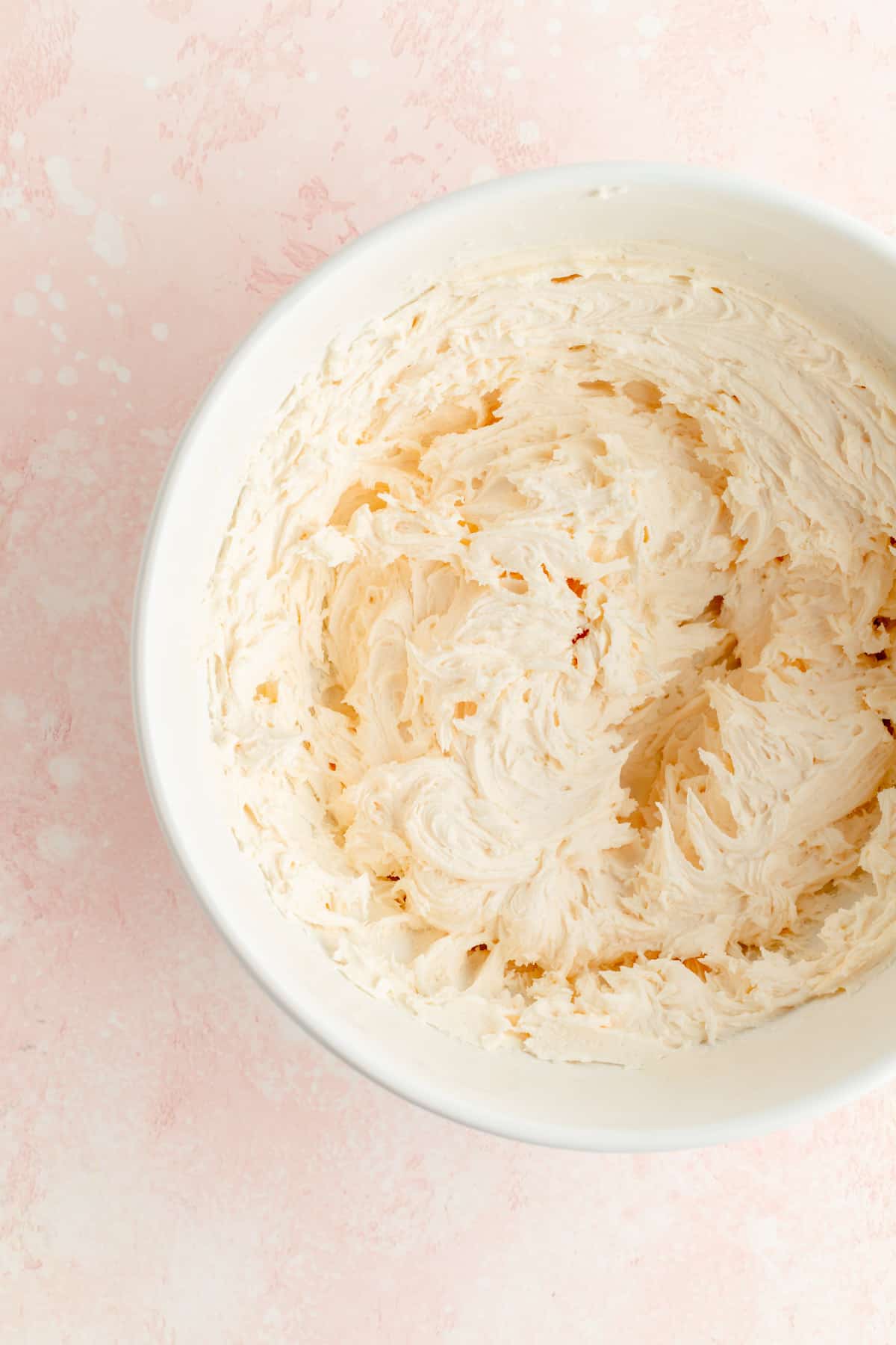 Overhead of whipped cream cheese frosting in white bowl and pink background.