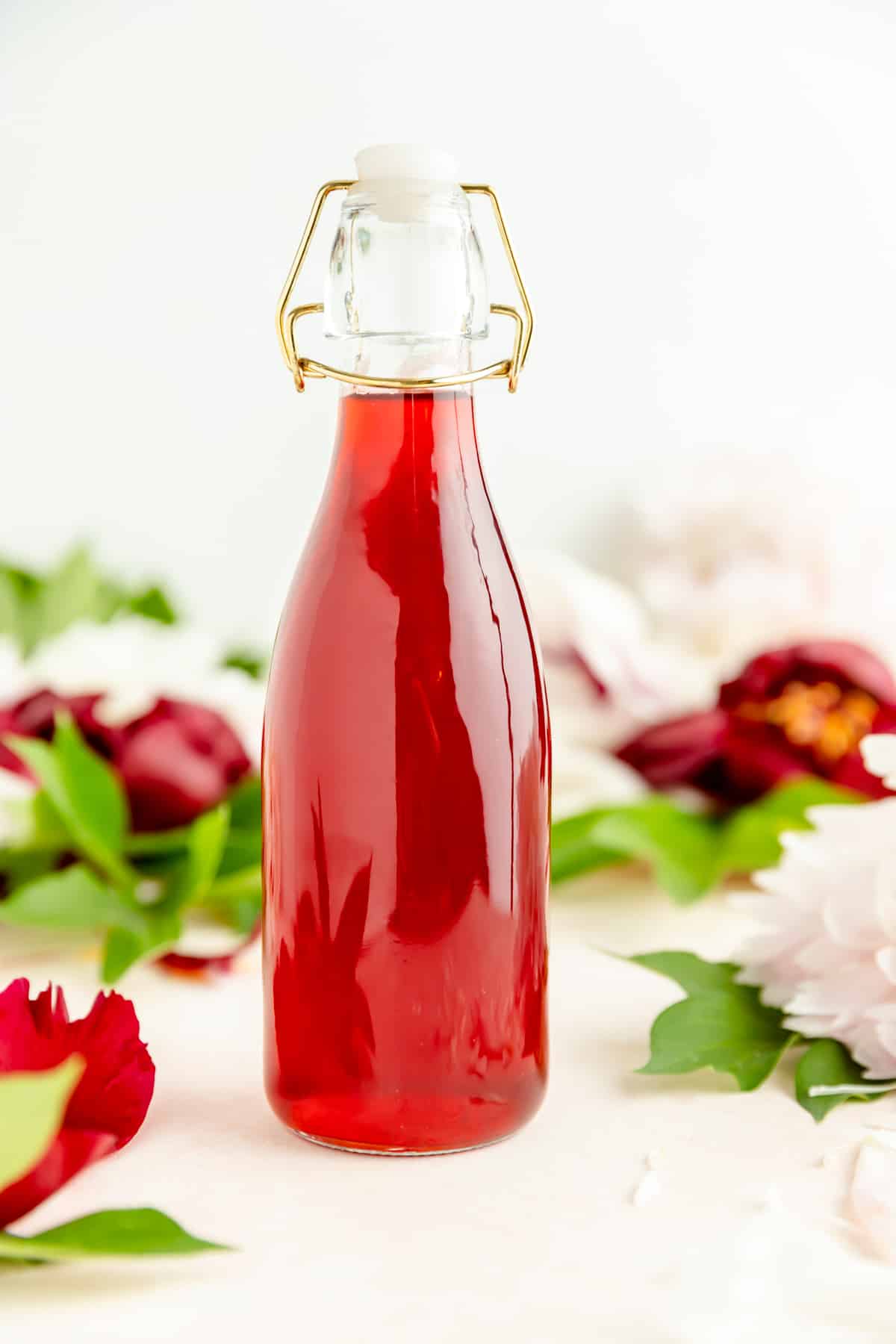 Close-up of red strawberry syrup in glass bottle with flowers on a white table.