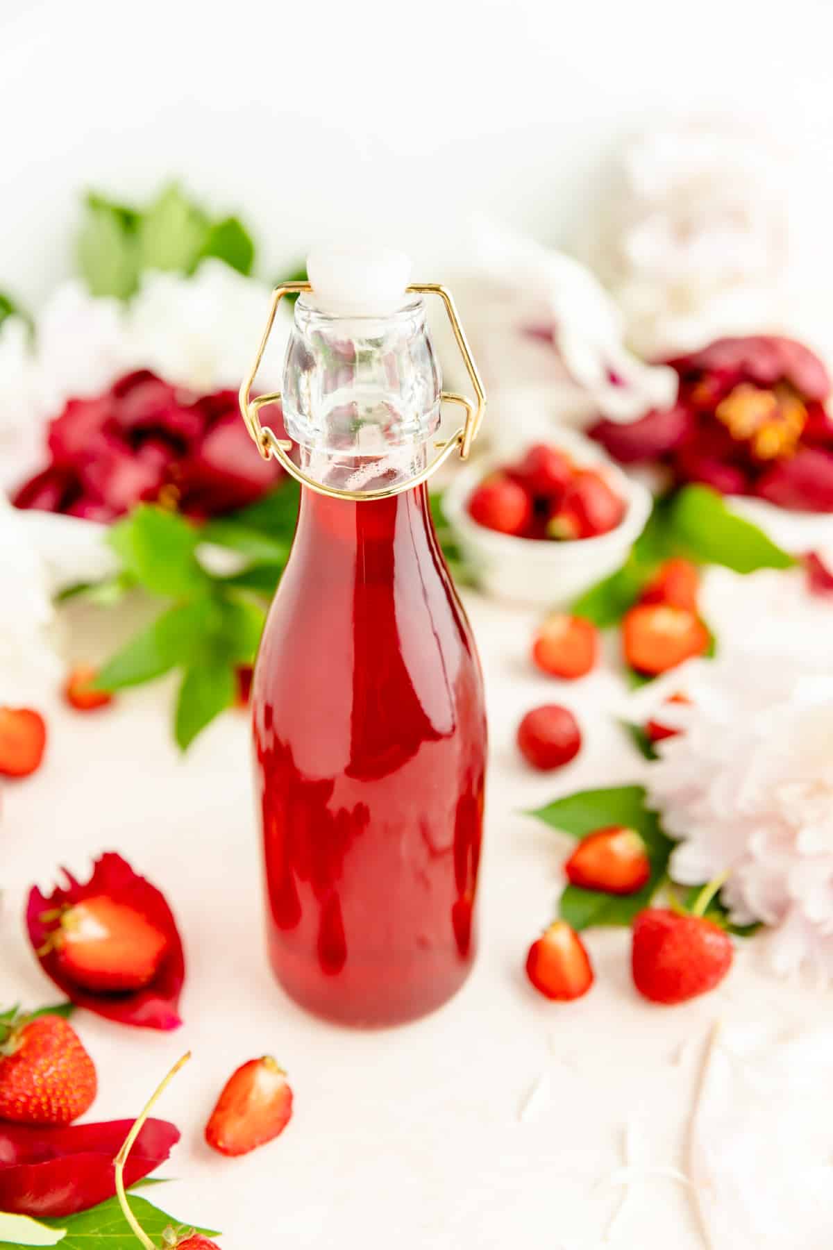 Close-up of red strawberry syrup in glass bottle with flowers and berries on table.