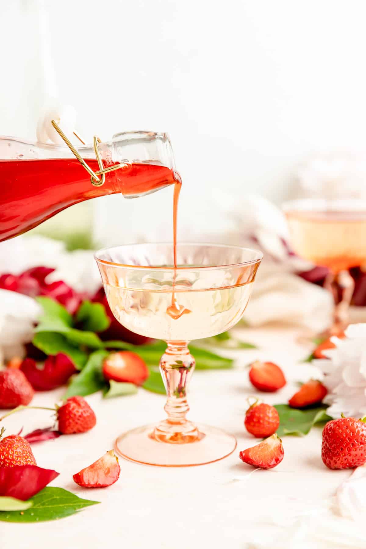 Glass bottle of strawberry syrup being poured into pick cocktail glass filled with Prosecco.