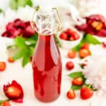 Close-up of red strawberry syrup in glass bottle with flowers and berries on table.