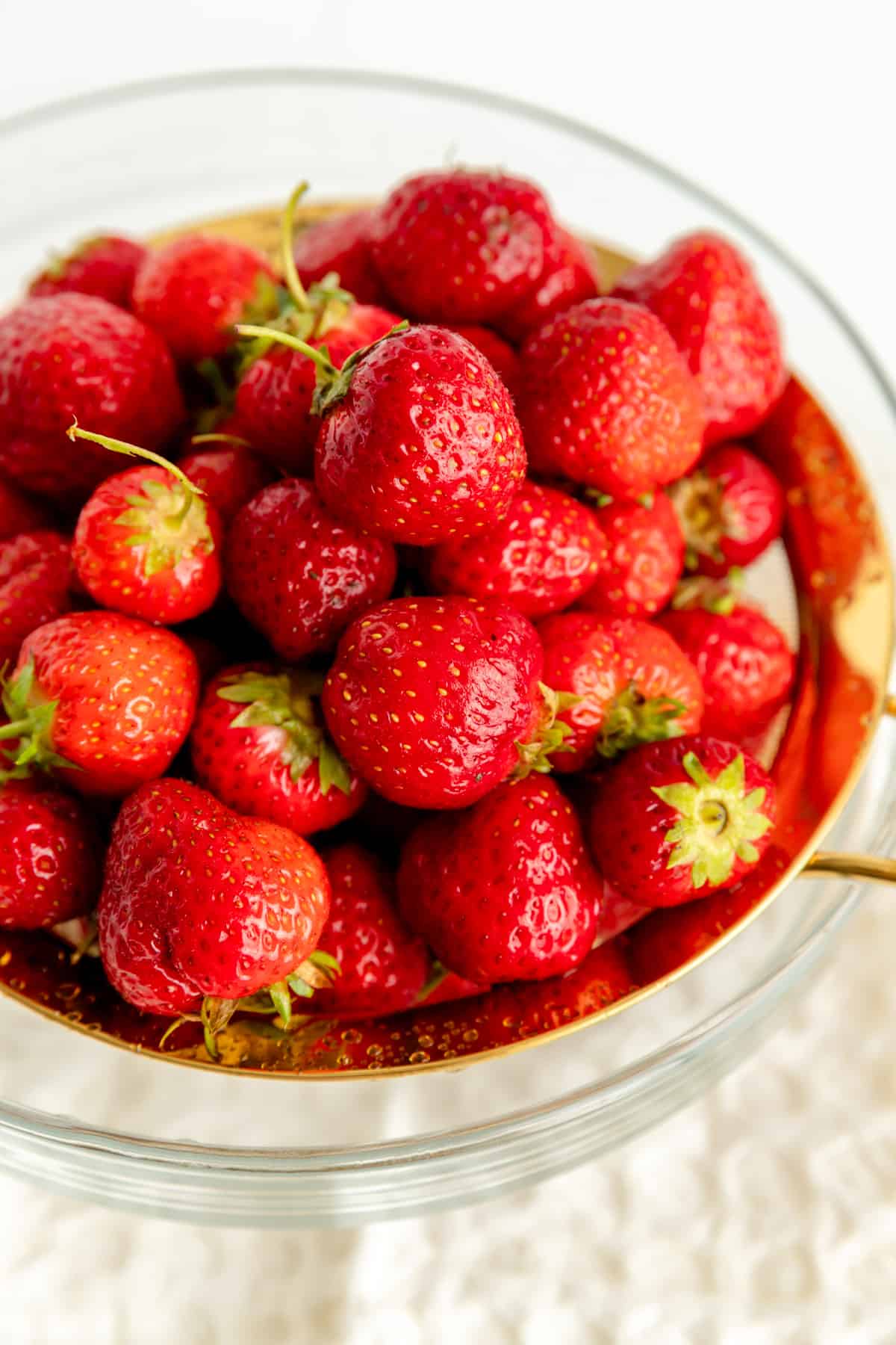 Fresh strawberries piled in a gold strainer over a glass bowl after being washed.