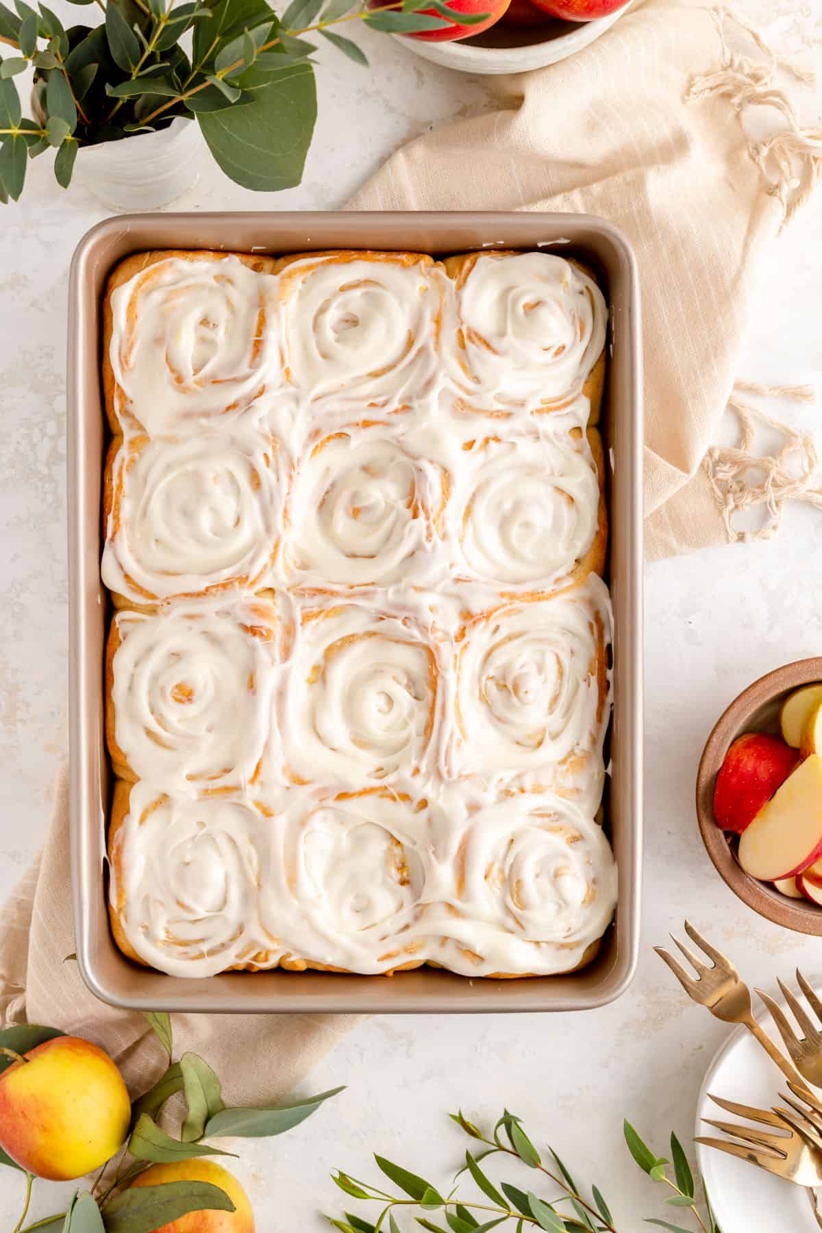 overhead of 12 iced cinnamon rolls in a champagne pan surrounded by apples and greens.