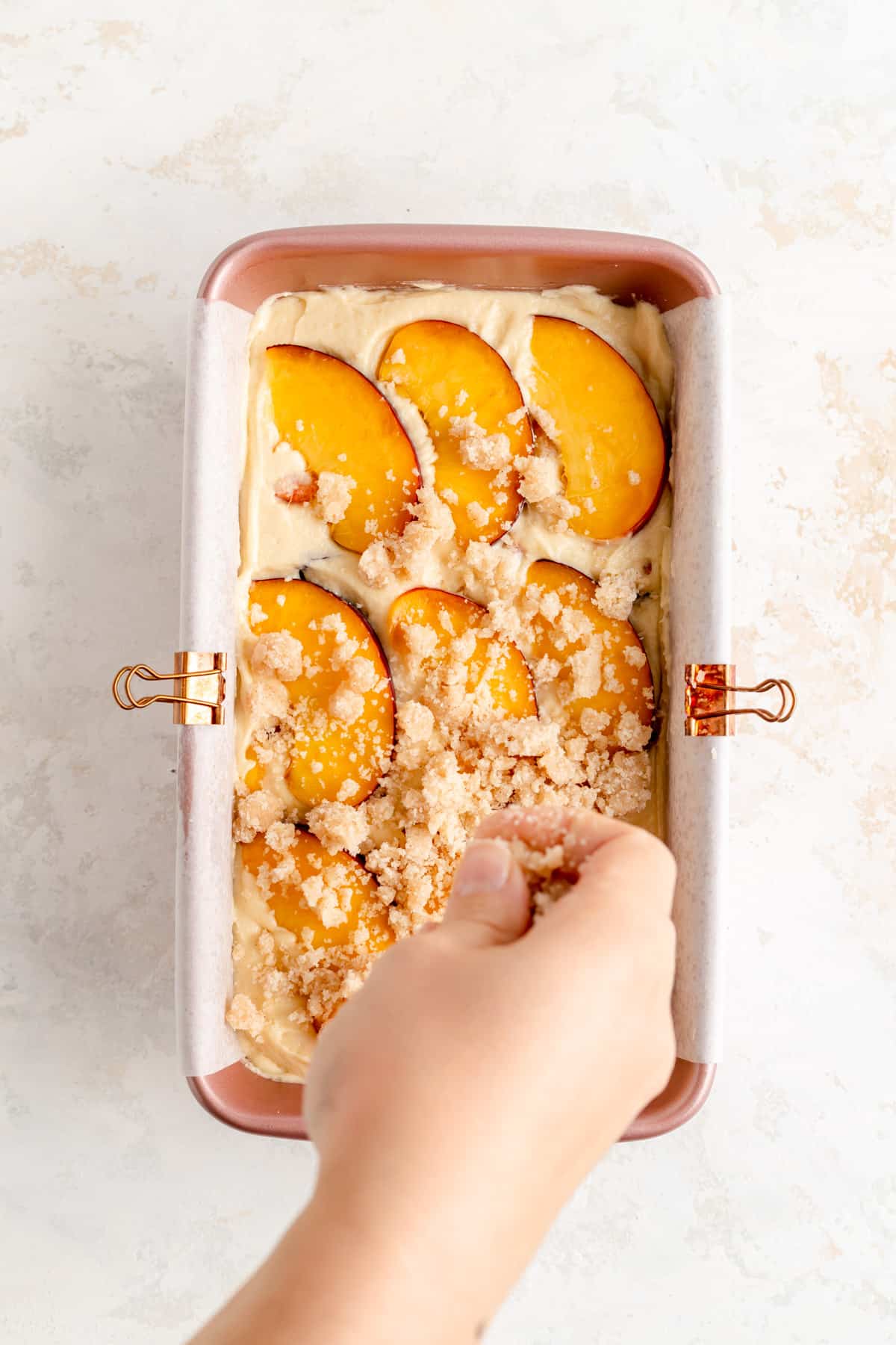 Hand sprinkling streusel topping over raw peach pound cake batter in pink loaf pan.