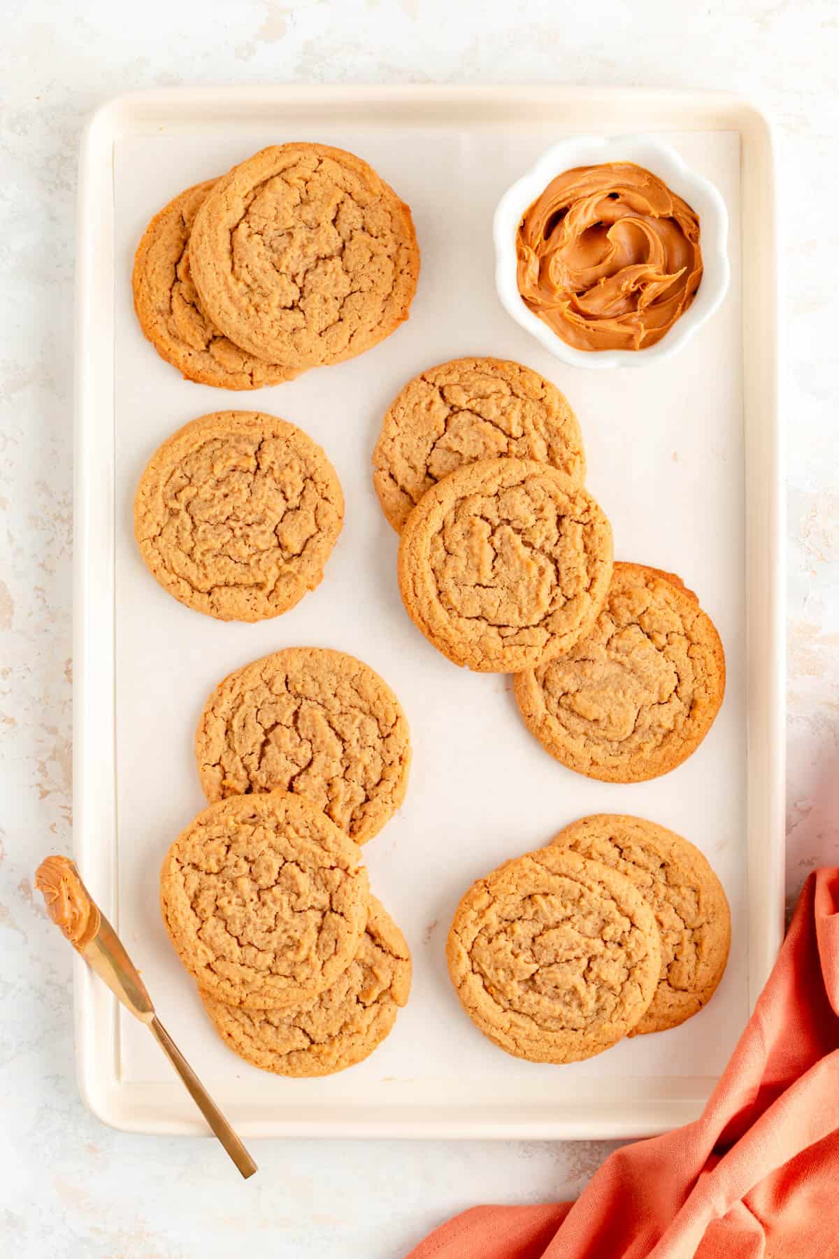 plain peanut butter cookies scattered on a white baking sheet with bowl of peanut butter.