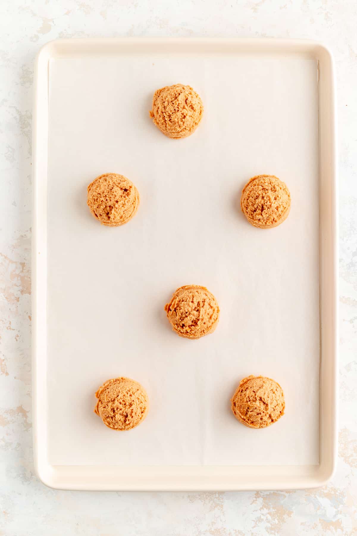 six plain peanut butter cookie dough scoops space on a lined white baking sheet.