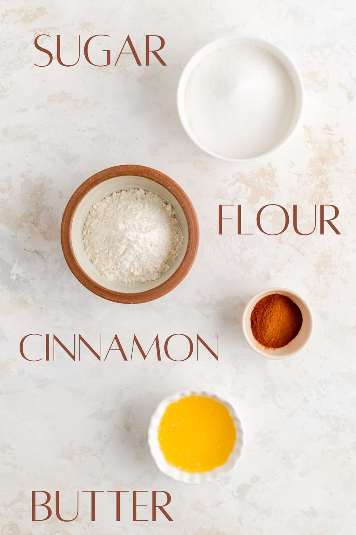 Labeled sugar flour cinnamon and butter in individual bowls on white plaster background.