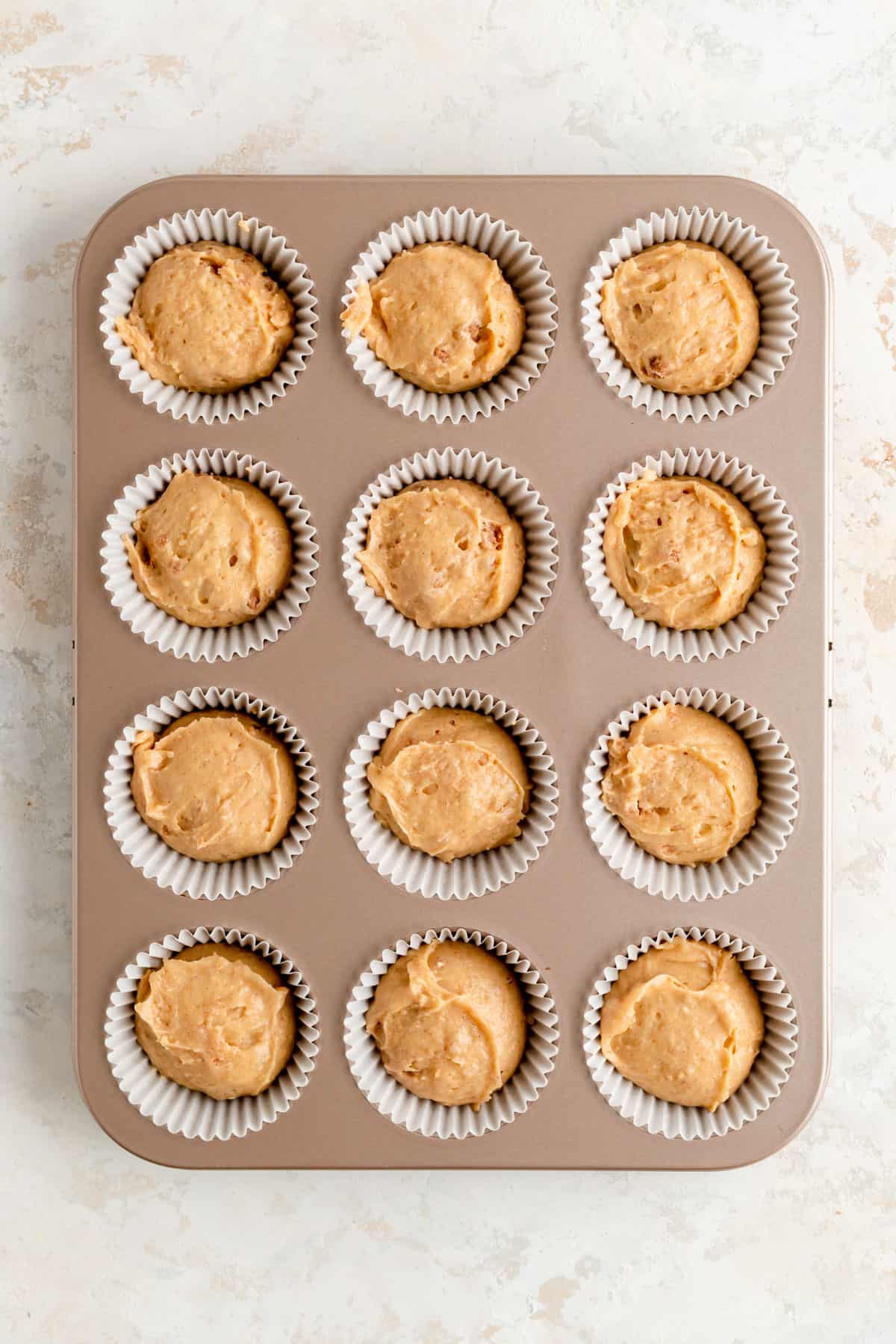 12 scoops of muffin batter in white papers in champagne pan on white background.