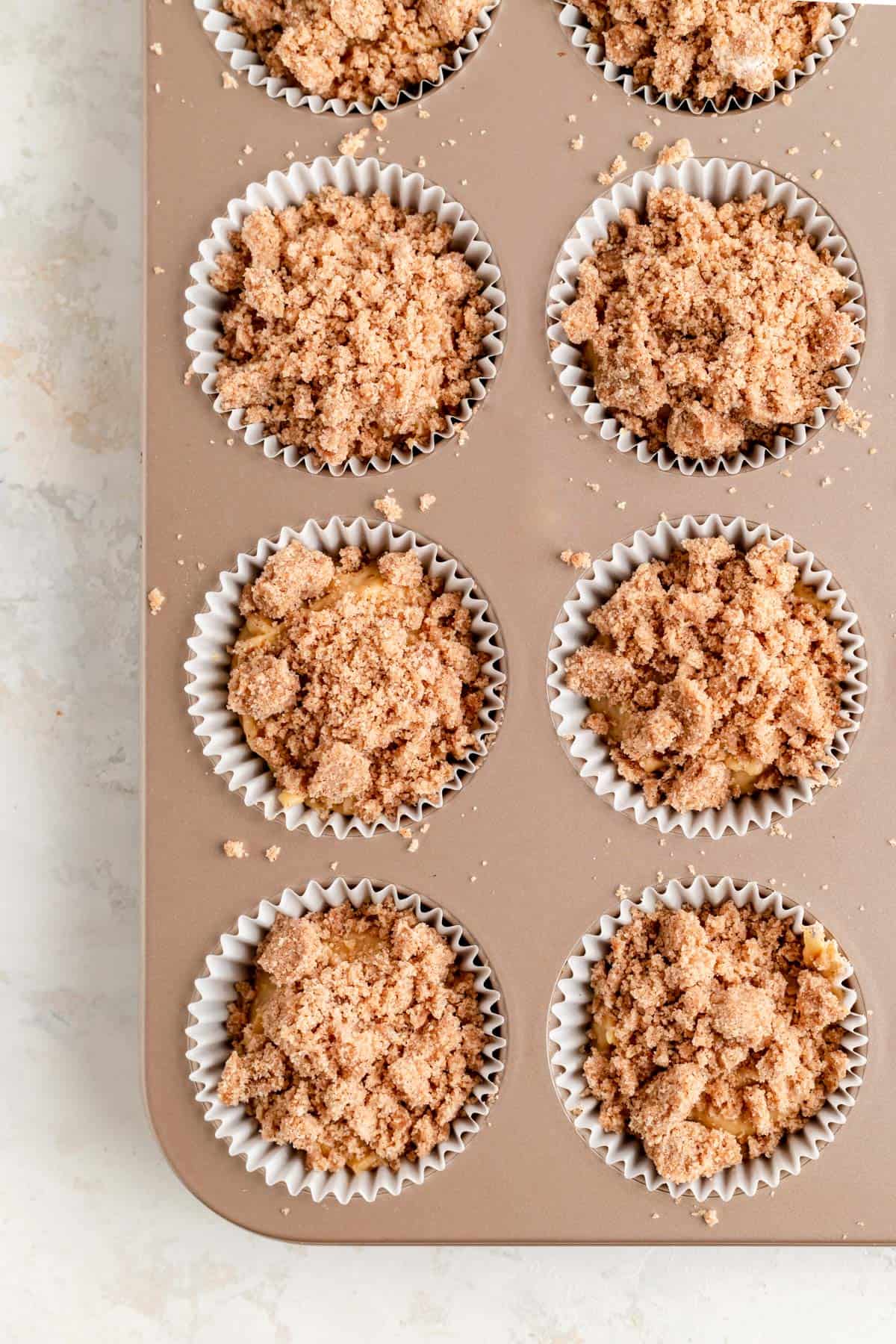 Overhead of raw batter and cinnamon streusel piled in muffin cups in champagne muffin pan.