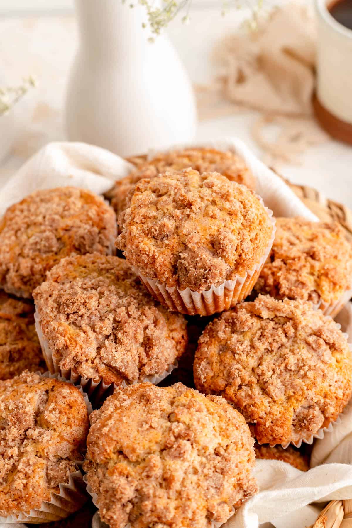 45° angle close up of stacked cinnamon streusel muffins with white vase and flowers behind.
