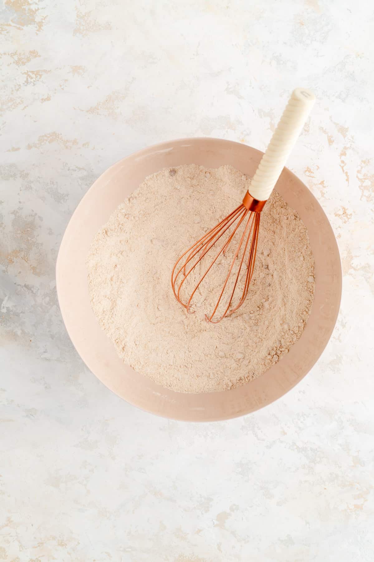 Mixed dry ingredients in tan bowl with copper and white whisk on white background.