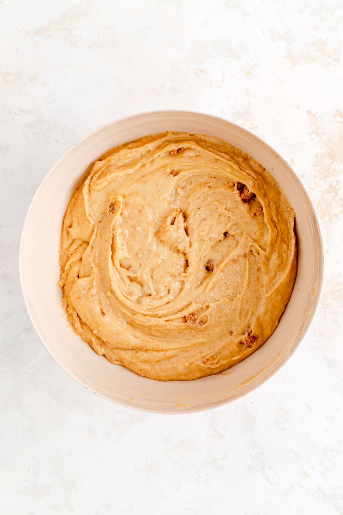 Final raw cinnamon muffin batter in tan bowl on white plaster background.