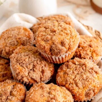 45° angle close up of stacked cinnamon streusel muffins with white vase and flowers behind.