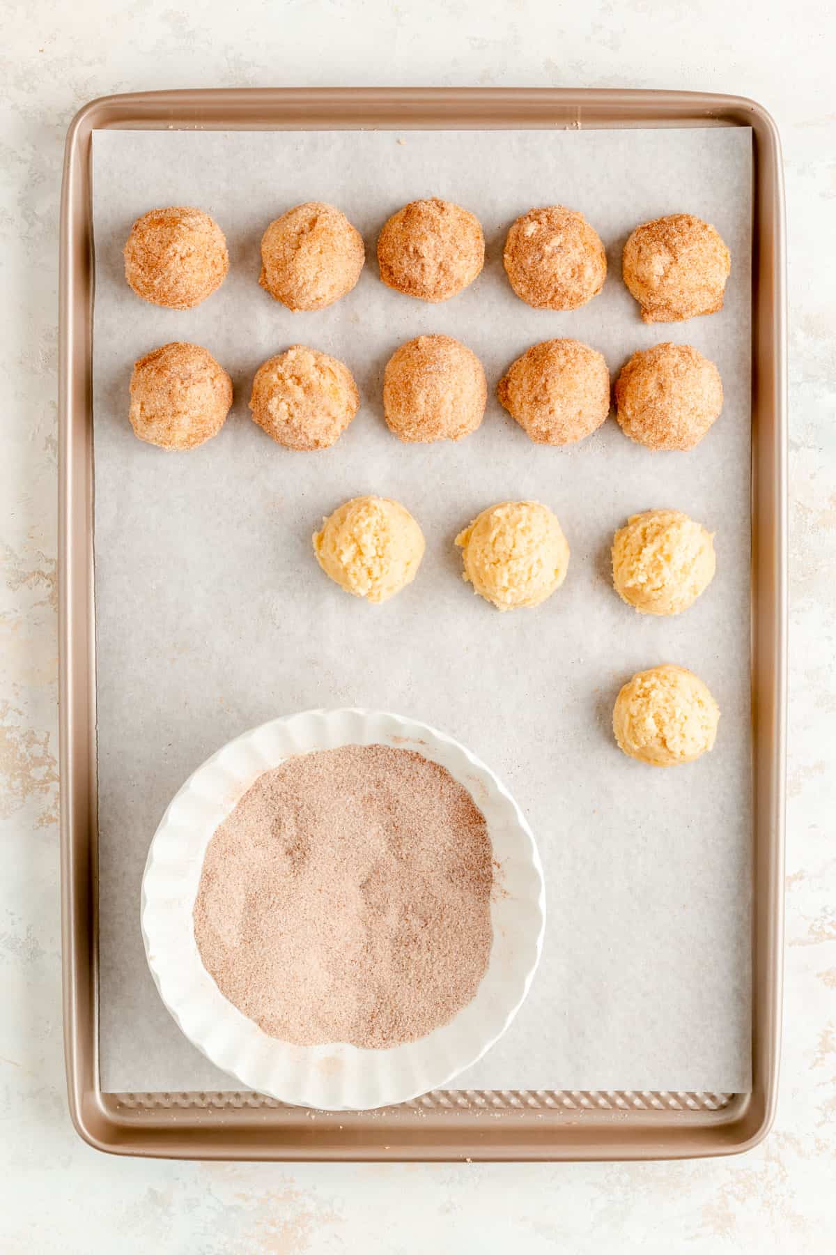 overhead of tan baking sheet with plain and coated dough balls and bowl of cinnamon sugar.