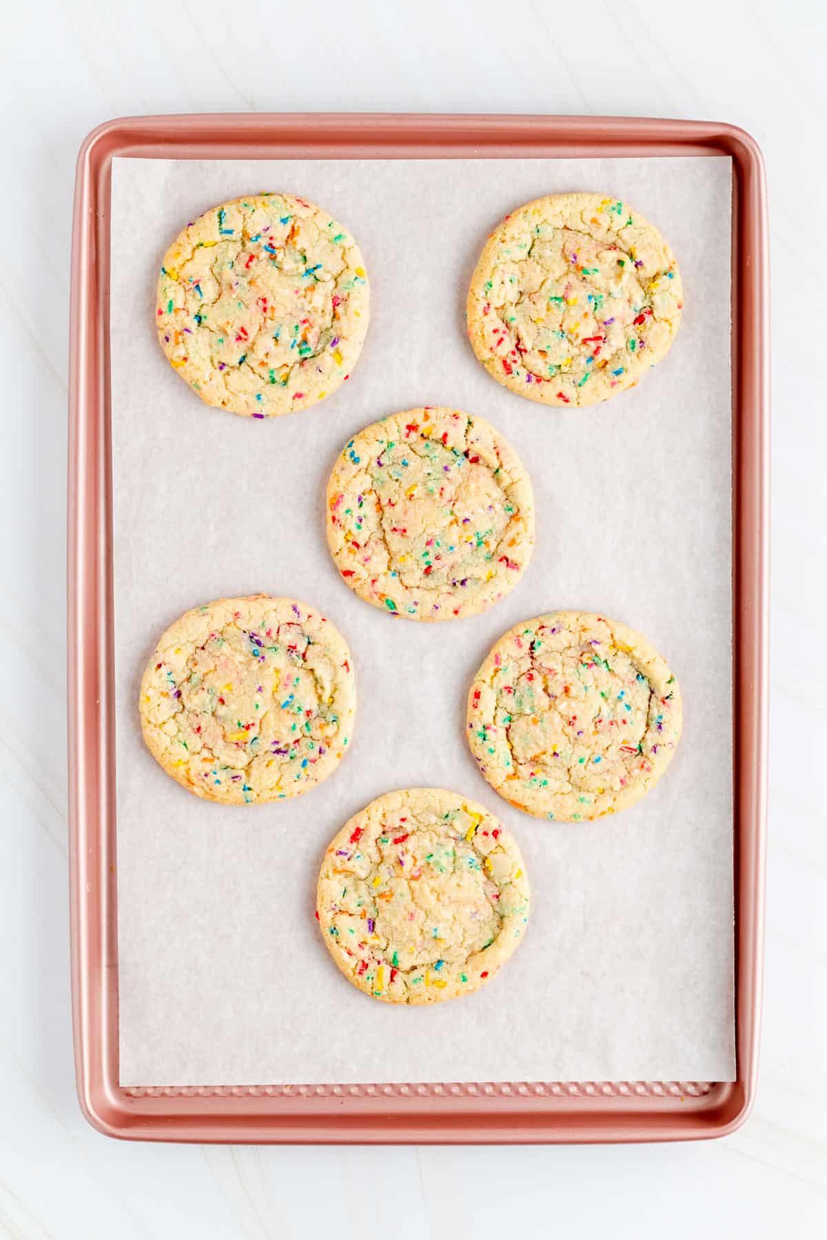Six baked funfetti cookies dough balls on a parchment lined pink baking pan.